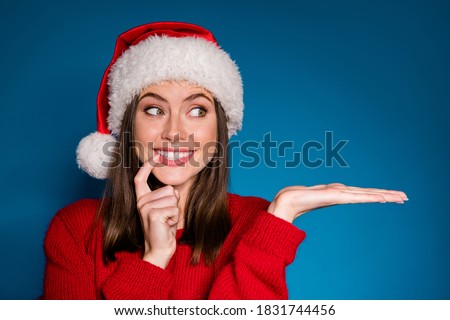Photo positive girl in santa claus hat hold hand demonstrate x-mas newyear advert promo touch finger teeth wear warm winter season clothes isolated blue gradient color background Royalty-Free Stock Photo #1831744456