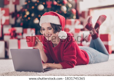 Photo of young lady lay floor hold cup laptop look screen drink wear santa cap red pullover decorated living room indoors