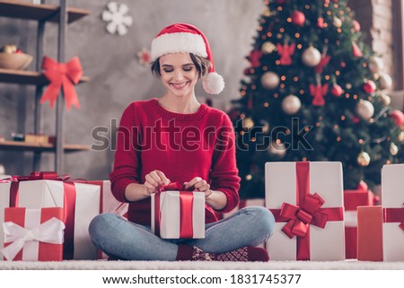 Photo of charming girl sit carpet unpack small giftbox wear santa headwear red sweater decorated living room indoors