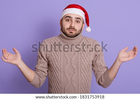 Christmas guy in Santa Hat and beige sweater showing helpless gesture with arm and hands, bearded handsome male making helpless sign, isolated on lilac background.