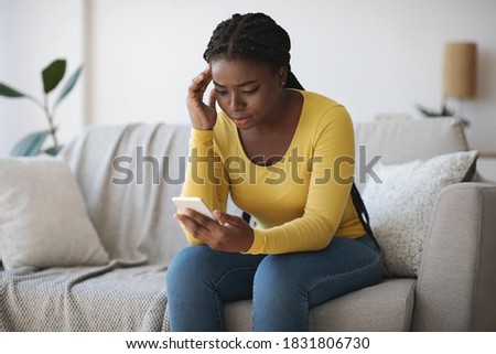 Worried black lady looking at smartphone screen, sitting on couch at home, concerned african american woman waiting for important call, reading unpleasant sms, spending time with phone in living room Royalty-Free Stock Photo #1831806730