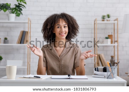Webinar, lecture, blog and lesson online. Happy african american woman gesturing and looking at camera, sitting at table with cup, notepad and pen in living room interior during pandemic, copy space