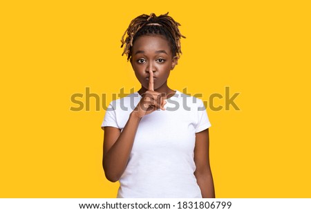 Pretty silent black woman making hush gesture, yellow studio background with copy space. Attractive african american young lady showing silence gesture, touching her lips with one finger