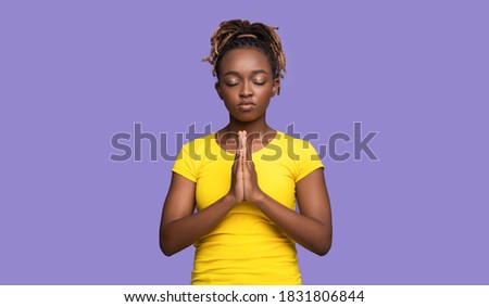 Young black woman showing clasped hands in pleading gesture, pretty please on purple studio background, copy space. Religious african lady praying with closed eyes, having hope for better future