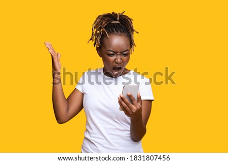 Furious black lady standing on yellow studio background, screaming at smartphone in her hand, gesturing, expressing anger, panorama with empty space. Unhappy african american lady holding mobile phone