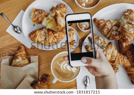 
A woman's hand on the phone takes pictures of a beautiful breakfast in a cafe - croissants and cappuccino with cinnamon