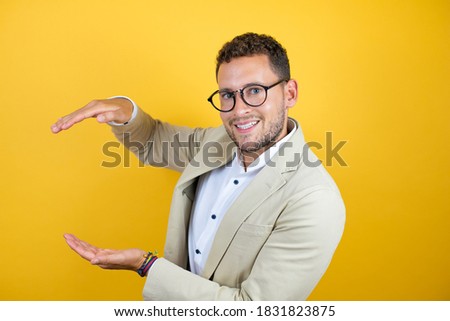 Young handsome businessman wearing suit over isolated yellow background surprised and gesturing with hands showing big and large size sign, measure symbol