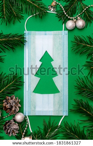 Medical protective mask with a cut-out silhouette of a Christmas tree on a green background.Frame of fir tree branches around the mask.Christmas and Happy New Year concept, coronavirus concept.
