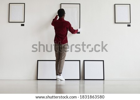 Full length back view at African-American man hanging blank frames on wall while planning art gallery or exhibition, copy space Royalty-Free Stock Photo #1831863580