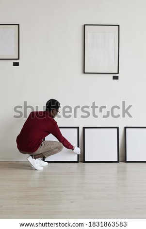 Vertical full length portrait of African-American man setting up blank black frames on floor while planning art gallery or exhibition, copy space