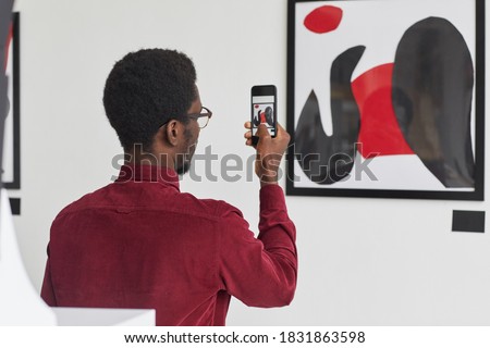 Back view portrait of young African-American man taking photo of painting via smartphone at contemporary art gallery exhibition, copy space