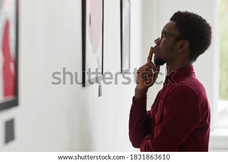 Side view portrait of young African-American man looking at paintings and thinking at art gallery or museum exhibition, copy space