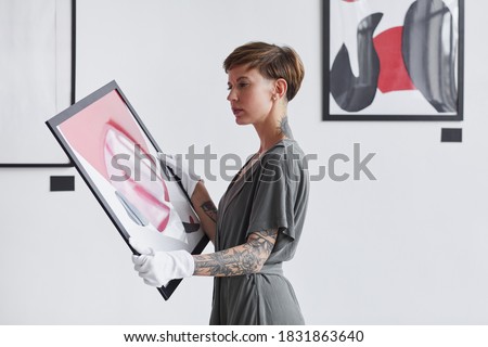 Waist up portrait of tattooed creative woman holding painting while planning art gallery exhibition, copy space