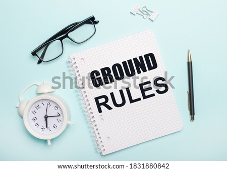 On a light blue background lie black glasses and a pen, a white alarm clock, white paper clips and a notebook with the words RULES OF THE EARTH. Business concept