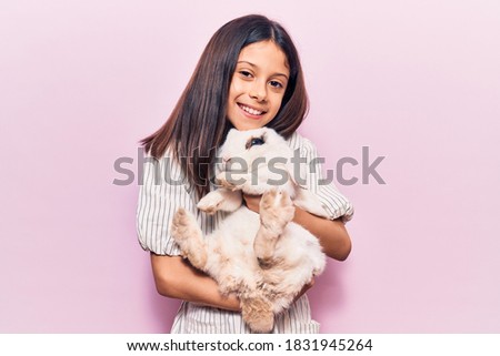 Adorable hispanic kid girl smiling happy. Holding cute rabbit standing over isolated pink background