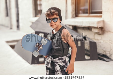 Side view of cool hipster boy in trendy outfit and sunglasses carrying skateboard and looking at camera, while standing on urban street and preparing for riding in summertime in city