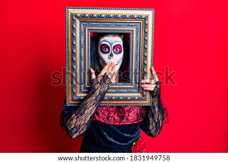 Young woman wearing day of the dead costume holding empty frame covering mouth with hand, shocked and afraid for mistake. surprised expression 