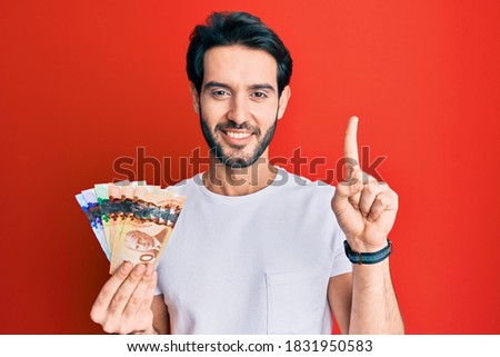 Young hispanic man holding canadian dollars smiling with an idea or question pointing finger with happy face, number one 