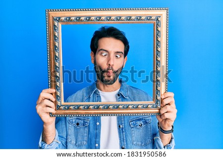 Young hispanic man holding empty frame making fish face with mouth and squinting eyes, crazy and comical. 