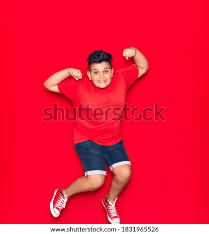 Adorable kid wearing casual clothes smiling happy. Jumping with smile on face doing strong gesture with arms over isolated red background