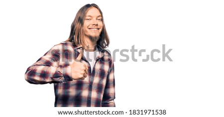 Handsome caucasian man with long hair wearing hipster shirt doing happy thumbs up gesture with hand. approving expression looking at the camera showing success. 