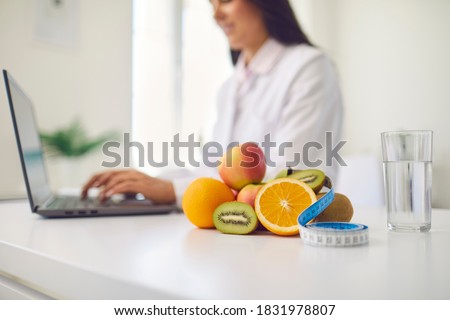 Close-up of fresh apples, kiwis and oranges with measuring tape and glass of clean water on office desk against blurred dietitian with laptop giving online weight loss and healthy diet consultation Royalty-Free Stock Photo #1831978807