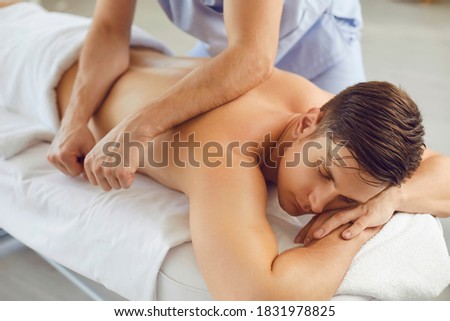 Young man enjoying professional body treatment in spa salon. Relaxed client lying on bed with eyes closed and getting remedial body massage done by male massagist in health center or massage room Royalty-Free Stock Photo #1831978825