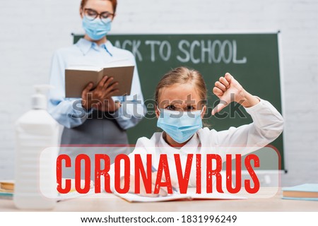 selective focus of schoolgirl in medical mask showing thumb down, teacher standing near chalkboard with book near coronavirus lettering