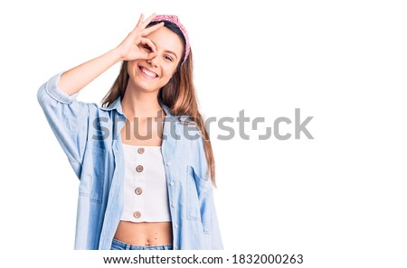 Young beautiful girl wearing casual shirt and diadem smiling happy doing ok sign with hand on eye looking through fingers 