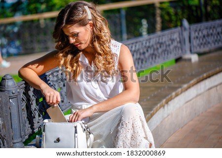 A young girl in a white dress sits in the park on a bench with a white handbag. Summer sunny day