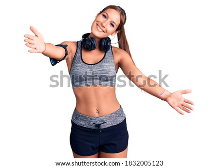 Young beautiful hispanic woman wearing gym clothes and using headphones looking at the camera smiling with open arms for hug. cheerful expression embracing happiness. 