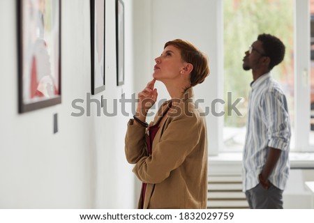 Side view portrait of elegant young woman looking at paintings while exploring modern art gallery exhibition, copy space