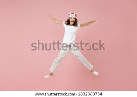 Full length portrait of laughing young woman in white pajamas home wear jumping spreading hands legs while resting at home isolated on pastel pink background studio. Relax good mood lifestyle concept