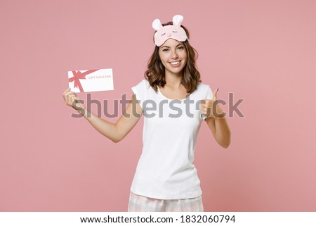 Smiling young woman 20s in pajamas home wear sleep mask showing thumb up holding gift certificate while resting at home isolated on pink background studio portrait. Relax good mood lifestyle concept