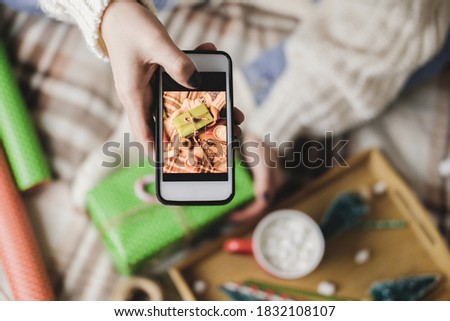 Female hand with smartphone takes pictures of New Year's or Christmas flatlay for social networks. Creating content. Wooden tray with cocoa mug marshmallows, toy tree, candle, stars, wrapped gift.