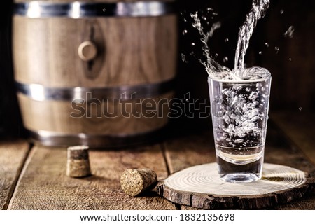 Pouring distilled alcohol into a shot glass, drops flying, served on wooden planks. Vintage bench with oak barrel and wooden stopper around Royalty-Free Stock Photo #1832135698