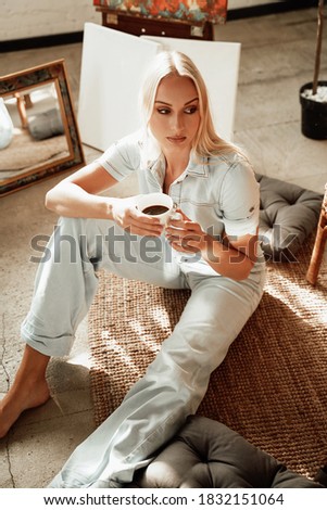 Blond and graceful female artist resting after work around pillow on carpet and drinking coffee in warm cosy room in the morning.