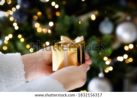 Close up of hand holding a golden gift box with Christmas tree and lights on the background, present and Christmas holiday concept sparkle background