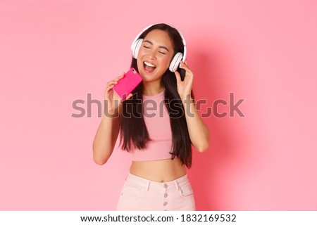 Waist-up portrait of attractive stylish asian girl close eyes and singing along song while listening music in headphones, playing karaoke game on mobile phone, standing over pink background Royalty-Free Stock Photo #1832169532