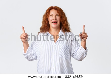 Wondered and amazed, upbeat middle-aged woman react to wonderful news, pointing and looking up with excited smile, reading banner, react to awesome promo offer, standing white background