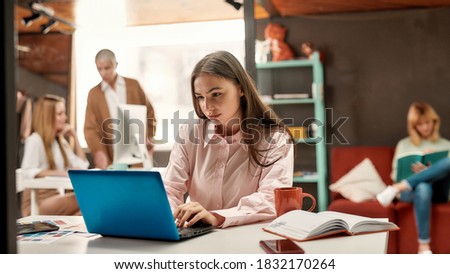 Creative female. A pretty yound brutette sitting at a table working with a laptop with her female colleagues discussing fashion trends on a background of a bright cosy designers workshop. Business