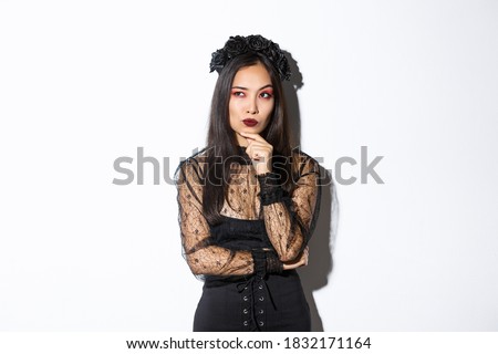 Thoughtful elegant evil witch in black lace dress, gothic wreath and makeup looking upper left corner. Woman making plans for halloween party, thinking over white background