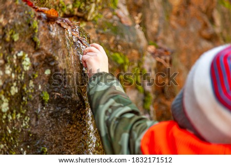 A curious caucasian boy wearing winter clothes is reaching out to a piece of icicle that formed over a tiny stick on side of a mossy rock. This is a cold day in forest.