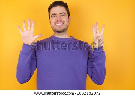 Handsome man with sweatshirt over isolated yellow background showing and pointing up with fingers number eight while smiling confident and happy.