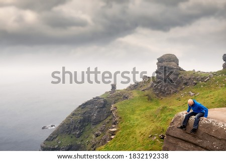 Bald man with beard in blue jacket sitting on an edge of a cliff and looking down, Cliff of Moher, county Clare, Ireland, Low cloudy sky.