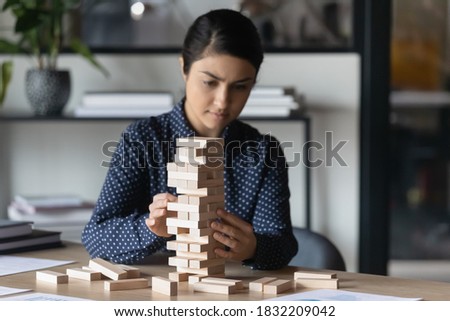 Pensive young 30s indian female employee sit at desk in office engaged in strategic thinking activity. Smart focused mixed race ethnicity woman brainstorm play wooden stack game alone at workplace.