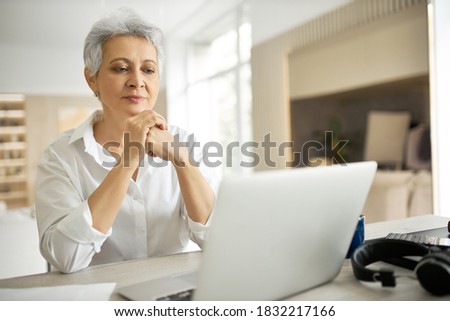 Electronic gadgets, devices and networking. Elegant gray haired mature female editor reading article for online magazine, sitting in front of open generic laptop, clasping hands, having focused look