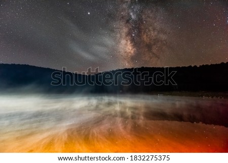 Night dark sky with milky way in Spruce Knob Lake in West Virginia with rising red yellow mist from water and reflection of stars landscape view
