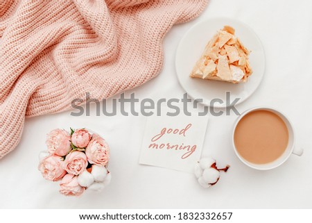 Cup of coffee with milk, piece of cake, rose flowers, pink knitted plaid or blanket, card with text GOOD MORNING. Breakfast in bed. Stylish home interior decor. Flat lay, top view.