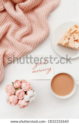 Cup of coffee with milk, piece of cake, rose flowers, pink knitted plaid or blanket, card with text GOOD MORNING. Breakfast in bed. Stylish home interior decor. Flat lay, top view.